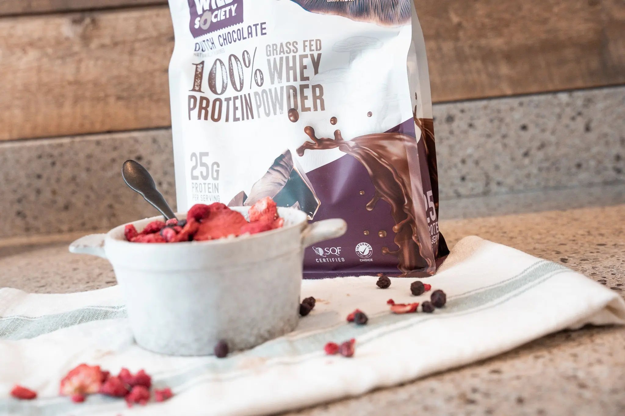 Full-sized package of Wild Society Grass-Fed Whey Protein Powder behind a bowl of protein oatmeal with dried berries on top.