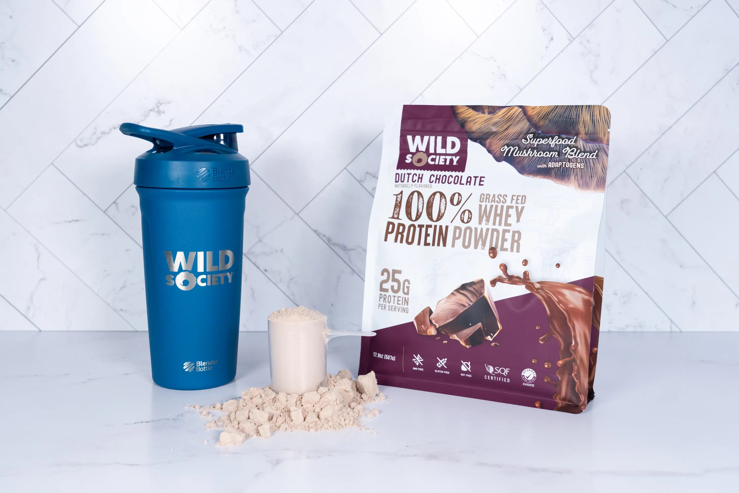 Full-size package of Wild Society Chocolate Whey Protein powder next to a shaker bottle and a scoop or protein powder for a smoothie.