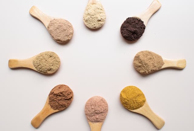 Eight wooden spoons with different colored low carb protein powders arranged in a circle