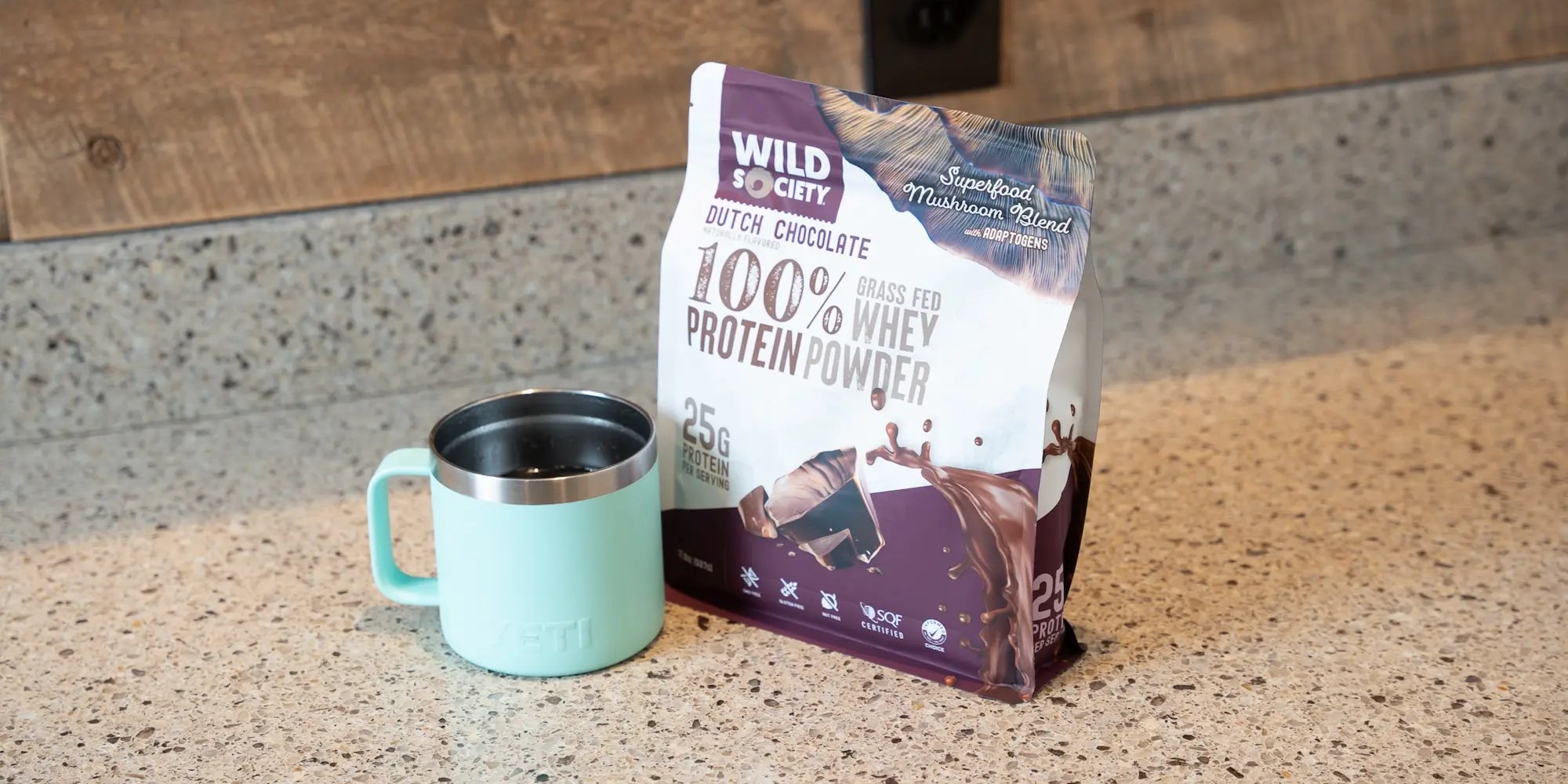 Full-size package of Wild Society Grass-Fed Whey Protein Powder next to a mug of hot coffee.