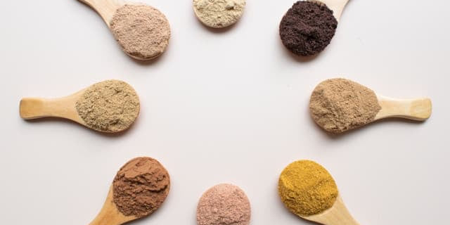 Eight wooden spoons with different colored low carb protein powders arranged in a circle