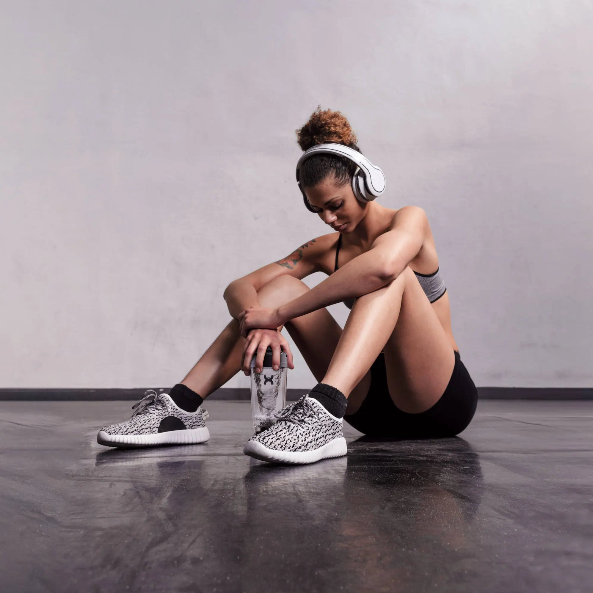 Person sitting on the floor wearing headphones and sneakers, with a glass of water in hand.